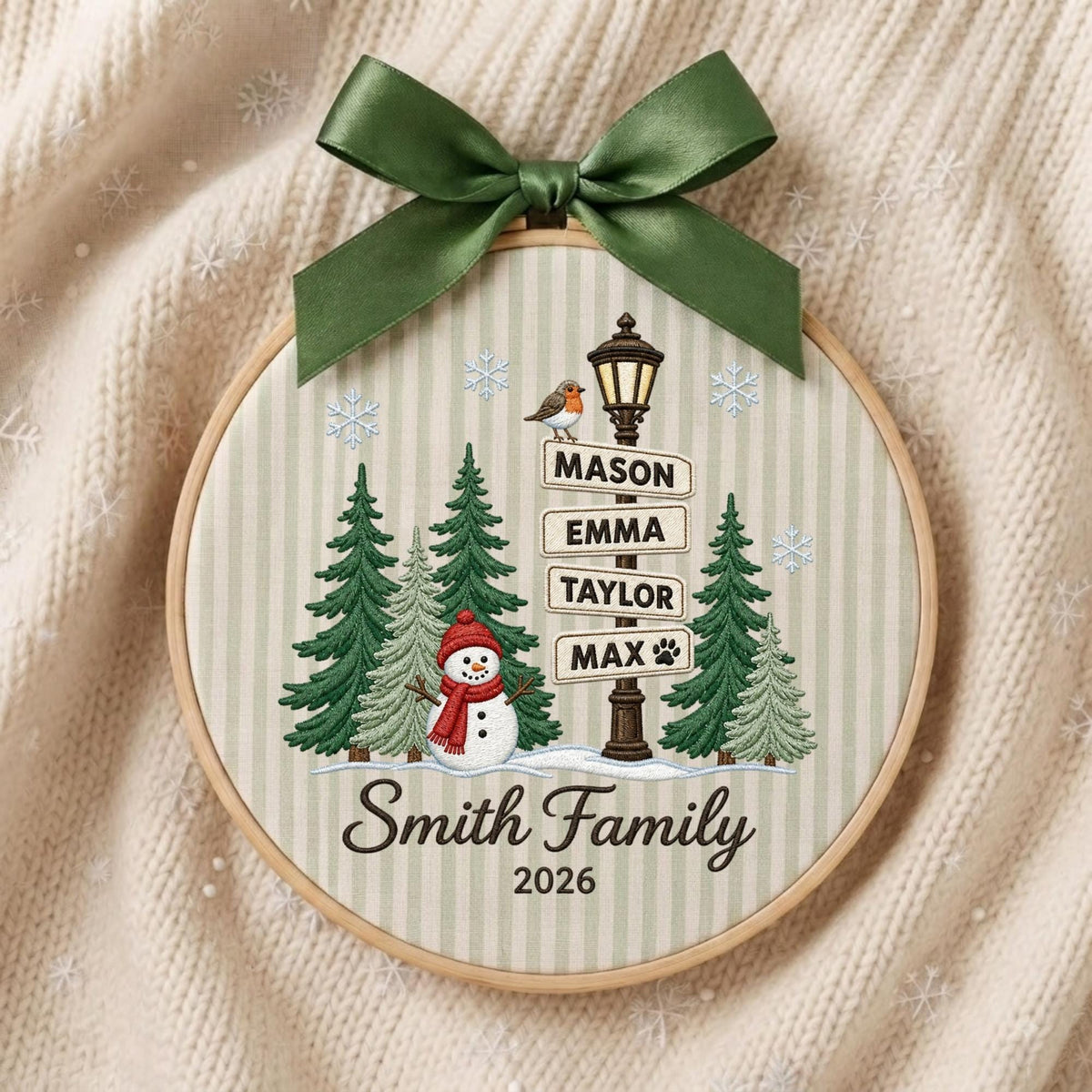 May include: A round embroidery hoop ornament featuring a light green and white striped fabric background. It displays a snowman, evergreen trees, a street lamp with signs reading Mason, Emma, Taylor, Max, and the text Smith Family 2026, topped with a green bow.