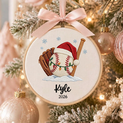 May include: A round embroidery hoop ornament featuring a baseball with a red Santa hat and string lights, a brown baseball glove, and a wooden bat. The design includes blue snowflakes, the name Kyle, and the year 2026, topped with a light pink ribbon.