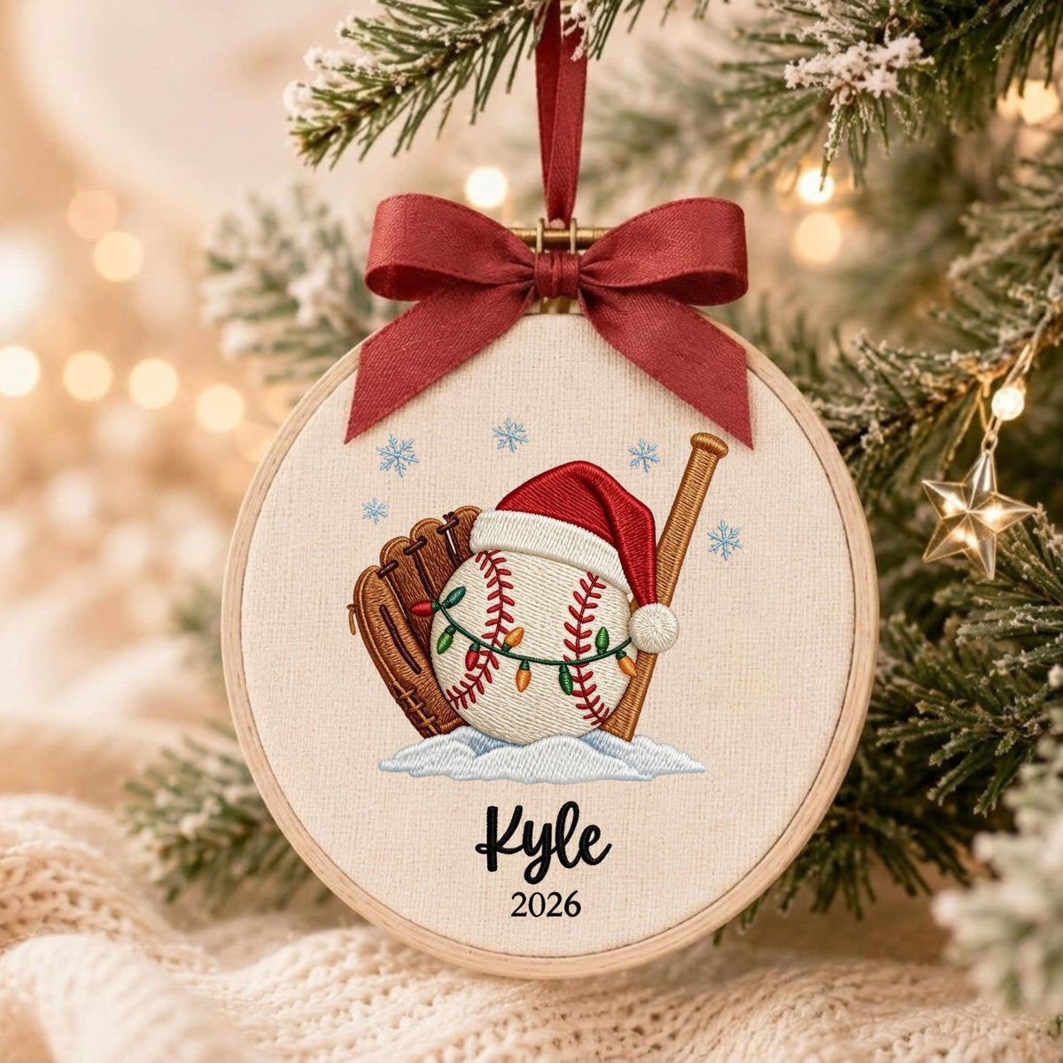 May include: A round wooden embroidery hoop ornament featuring a baseball, a brown leather glove, and a wooden bat. The baseball wears a red Santa hat and is wrapped in string lights. The text Kyle and 2026 is embroidered below. A red ribbon bow hangs from the top.