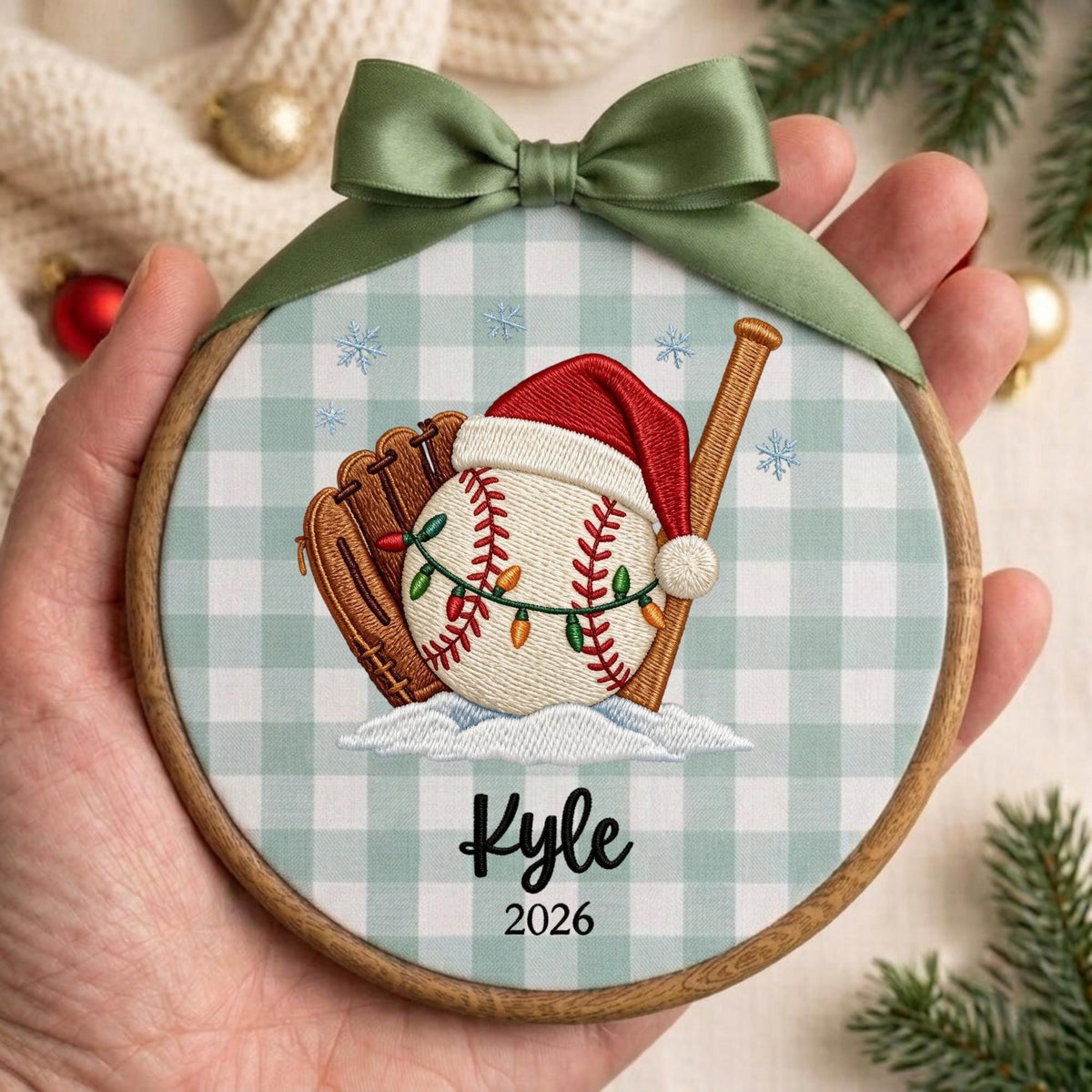 May include: A round wooden embroidery hoop ornament featuring a light green and white gingham fabric background. The design shows a baseball glove, a baseball wearing a Santa hat wrapped in string lights, and a wooden bat. Text reads Kyle 2026.