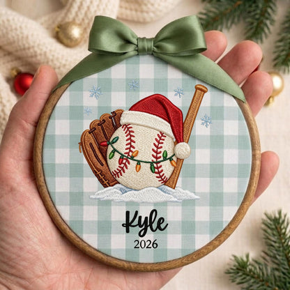 May include: A round wooden embroidery hoop ornament featuring a light green and white gingham fabric background. The design shows a baseball glove, a baseball wearing a Santa hat wrapped in string lights, and a wooden bat. Text reads Kyle 2026.