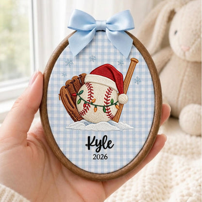 May include: An oval wooden embroidery hoop ornament featuring a light blue gingham fabric background. The embroidery depicts a baseball in a Santa hat, a glove, and a bat with Christmas lights. The text reads Kyle and 2026, topped with a light blue bow.