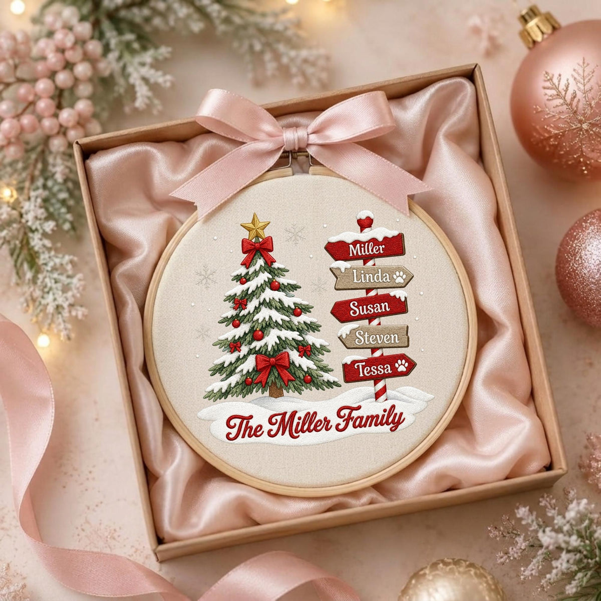 May include: A round embroidery hoop featuring a Christmas tree with red bows and a signpost with the names Miller, Linda, Susan, Steven, and Tessa, plus the text The Miller Family, topped with a light pink satin ribbon bow inside a gift box.