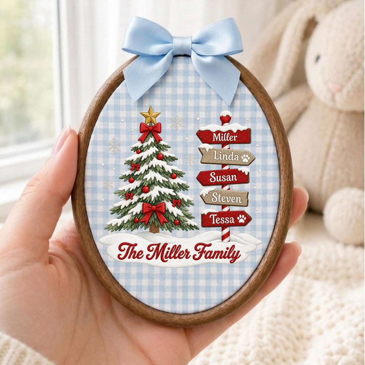 May include: An oval wooden embroidery hoop ornament featuring a light blue and white gingham fabric background. It displays a decorated Christmas tree and a signpost with the names Miller, Linda, Susan, Steven, and Tessa, plus The Miller Family text.