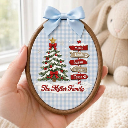 May include: An oval wooden embroidery hoop ornament featuring a light blue and white gingham fabric background. It displays a decorated Christmas tree and a signpost with the names Miller, Linda, Susan, Steven, and Tessa, plus The Miller Family text.