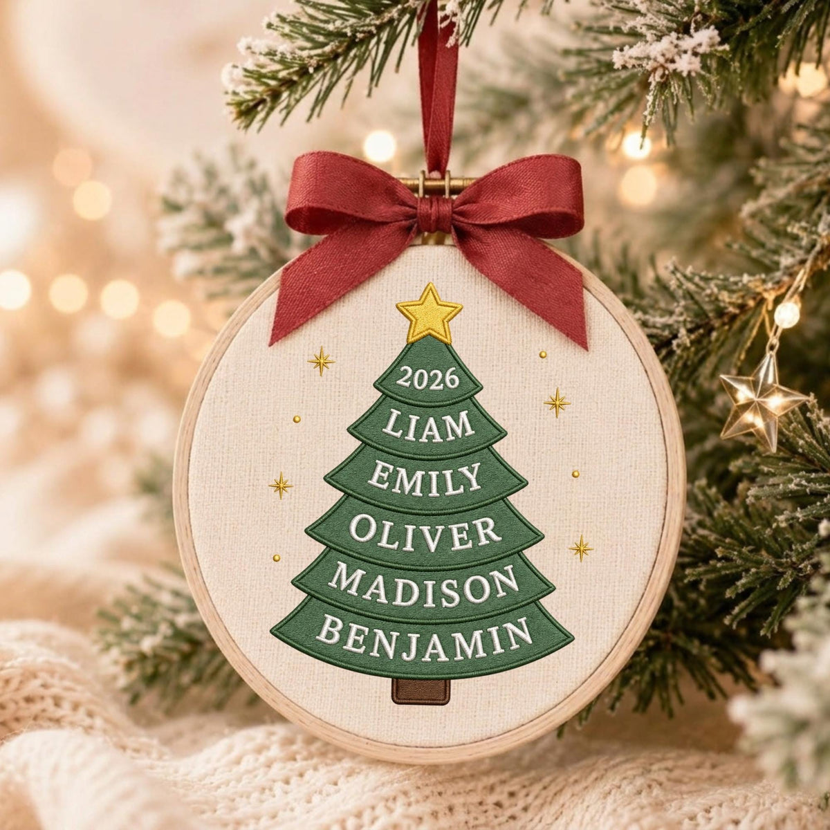 May include: A round wooden embroidery hoop ornament featuring a cream-colored fabric background. A green embroidered Christmas tree is centered with a yellow star on top. The text 2026, LIAM, EMILY, OLIVER, MADISON, BENJAMIN is stitched in white.