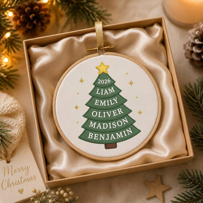 May include: A round embroidery hoop ornament featuring a white fabric background with a green embroidered Christmas tree. The tree displays the year 2026 and the names Liam, Emily, Oliver, Madison, and Benjamin. A yellow star sits atop the tree.