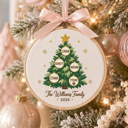 May include: A round wooden embroidery hoop ornament featuring a stitched green Christmas tree with a gold star. The tree displays five white circular ornaments with the names Logan, Scarlett, Nathan, Ethan, and Tyson. Text reads The Williams Family 2026.