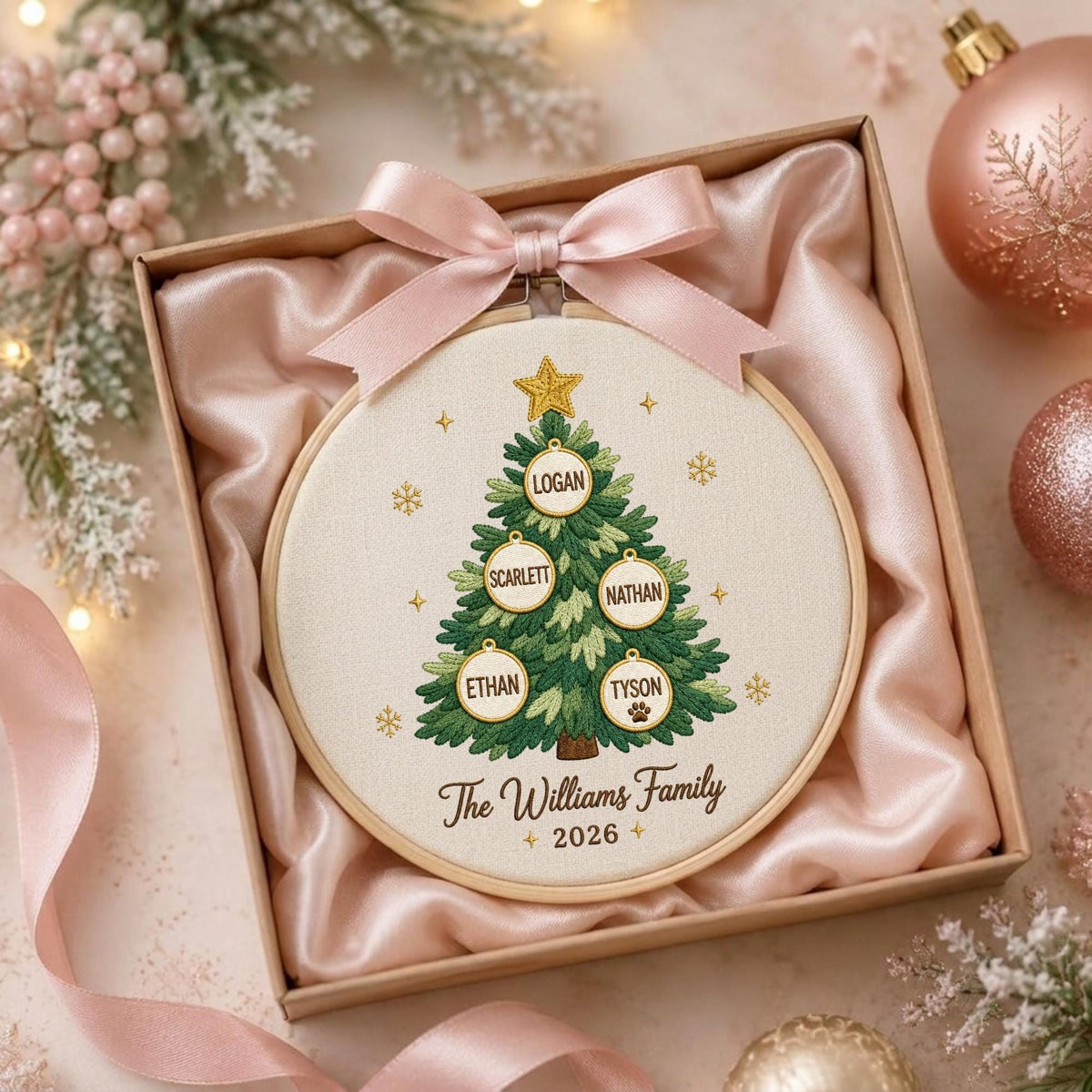 May include: A round wooden embroidery hoop containing a cream-colored fabric with a green Christmas tree design. The tree features five white circular ornaments with the names Logan, Scarlett, Nathan, Ethan, and Tyson, plus the text The Williams Family 2026.