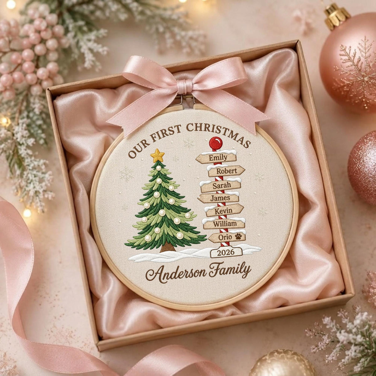 May include: A round wooden embroidery hoop containing fabric with a Christmas tree and a signpost featuring the names Emily, Robert, Sarah, James, Kevin, William, Orio, and the text Our First Christmas 2026 Anderson Family, topped with a pink ribbon.