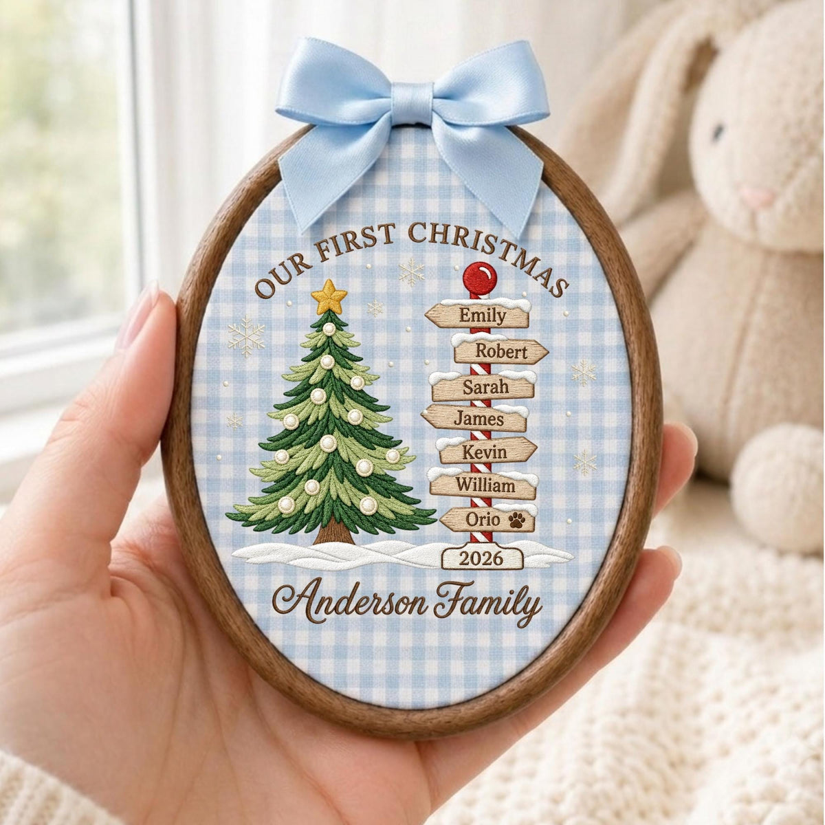 May include: An oval wooden embroidery hoop ornament featuring a light blue gingham fabric background. It displays a green Christmas tree with a yellow star, a signpost with names, and the text Our First Christmas 2026 Anderson Family. A light blue satin bow is attached to the top.