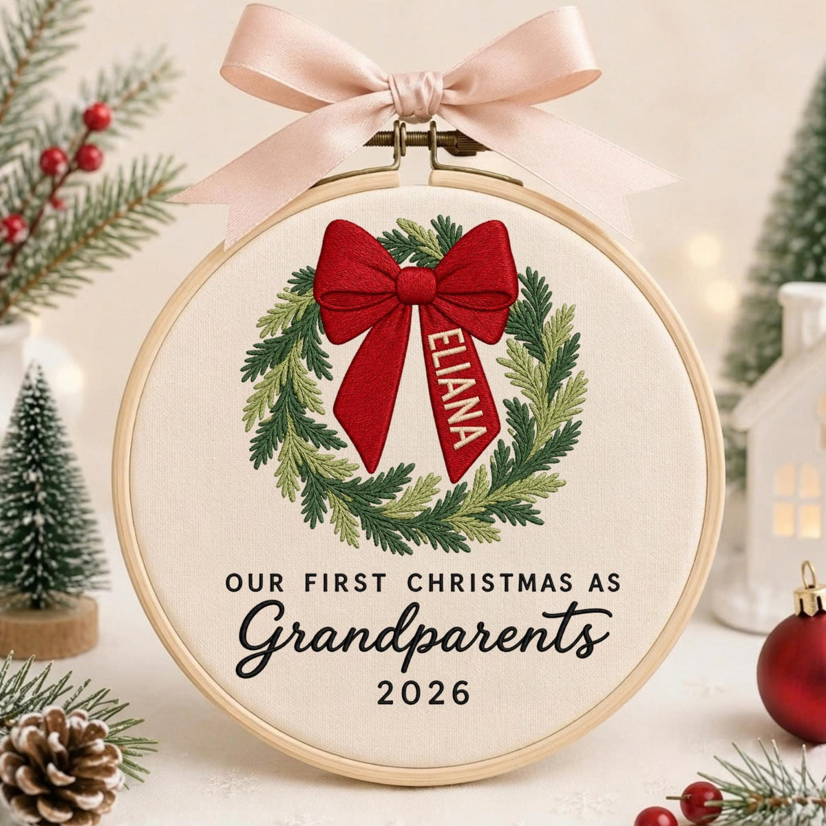 May include: A round wooden embroidery hoop containing off-white fabric with a green wreath, a red bow, and the name ELIANA embroidered on the ribbon. The text reads OUR FIRST CHRISTMAS AS Grandparents 2026. A light pink ribbon is tied at the top.