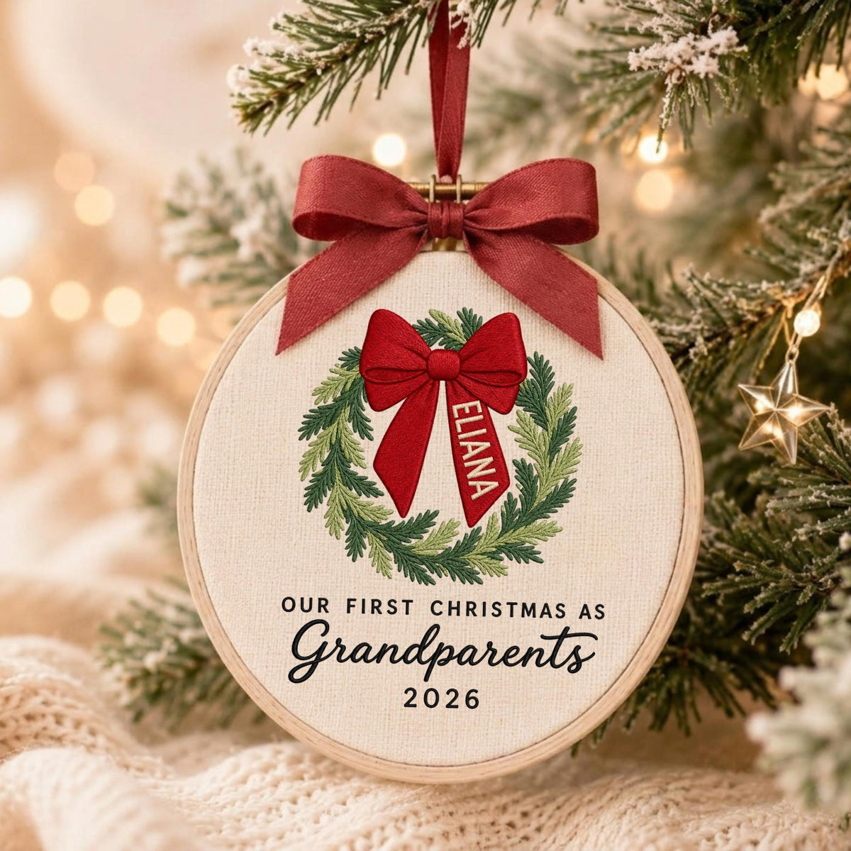 May include: A round wooden embroidery hoop ornament featuring a stitched green wreath with a red bow. The bow has the name ELIANA embroidered on one tail. Below the wreath, the text reads OUR FIRST CHRISTMAS AS Grandparents 2026. A red ribbon hangs from the top.