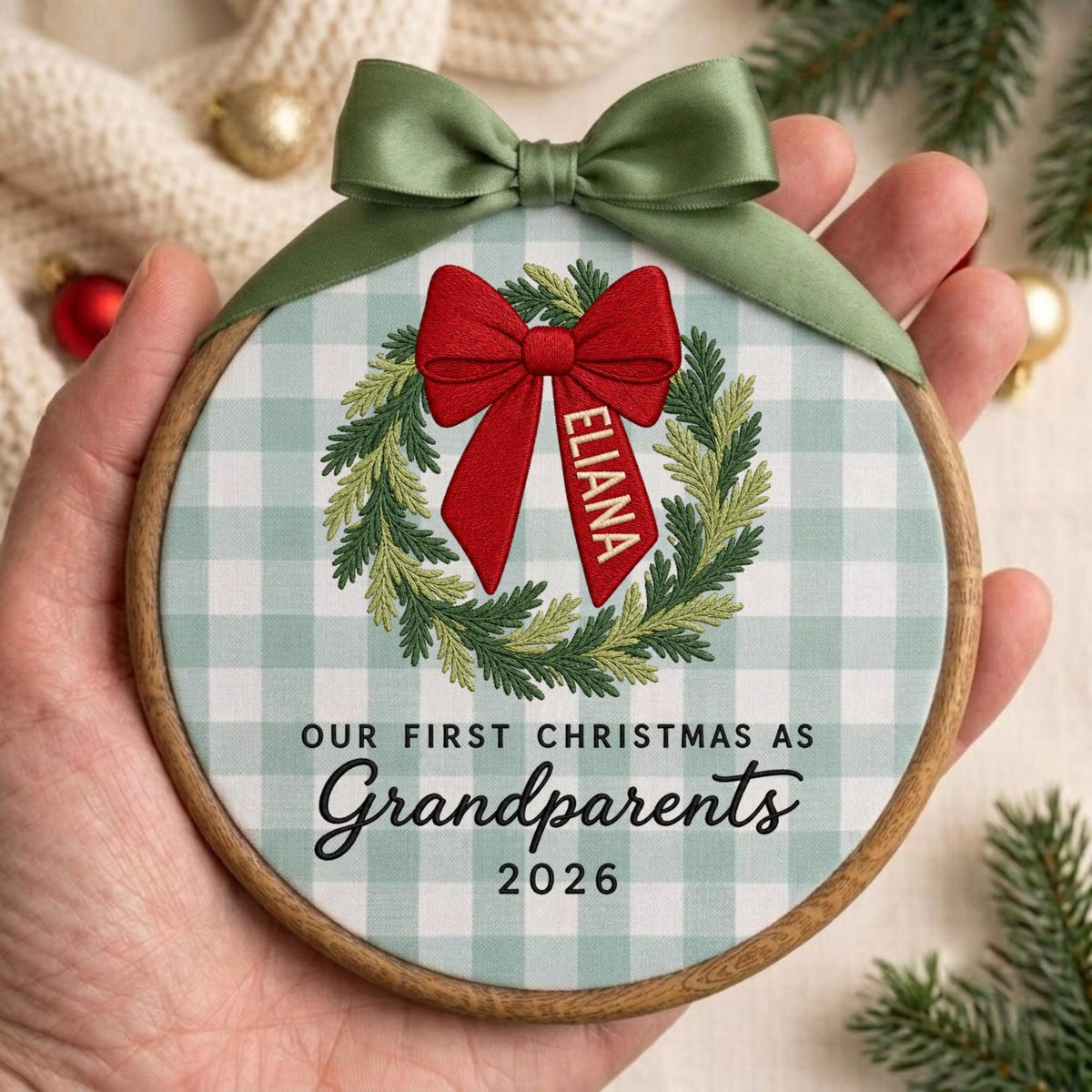 May include: A round wooden embroidery hoop ornament featuring a light green and white gingham fabric background. The design includes an embroidered green wreath with a large red bow, the name ELIANA, and the text OUR FIRST CHRISTMAS AS Grandparents 2026.