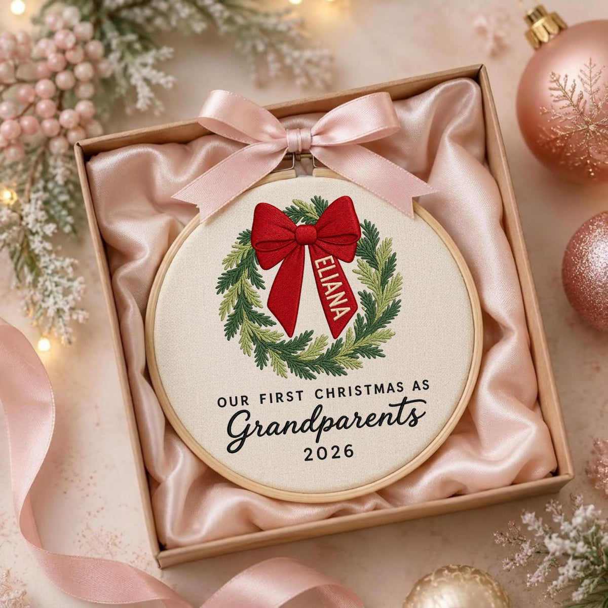 May include: A round wooden embroidery hoop containing white fabric with a stitched green wreath, a red bow, and the text "ELIANA", "OUR FIRST CHRISTMAS AS Grandparents 2026". A light pink satin ribbon is tied to the top of the hoop.