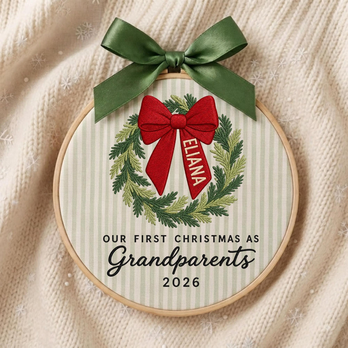 May include: A circular embroidery hoop featuring a light green and white striped fabric background. An embroidered green wreath surrounds a red bow with the name ELIANA. Below, black text reads OUR FIRST CHRISTMAS AS Grandparents 2026.