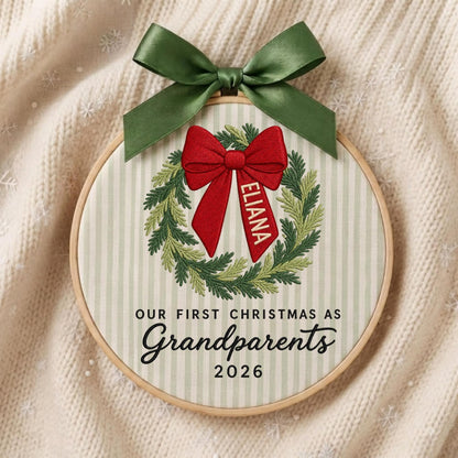 May include: A circular embroidery hoop featuring a light green and white striped fabric background. An embroidered green wreath surrounds a red bow with the name ELIANA. Below, black text reads OUR FIRST CHRISTMAS AS Grandparents 2026.