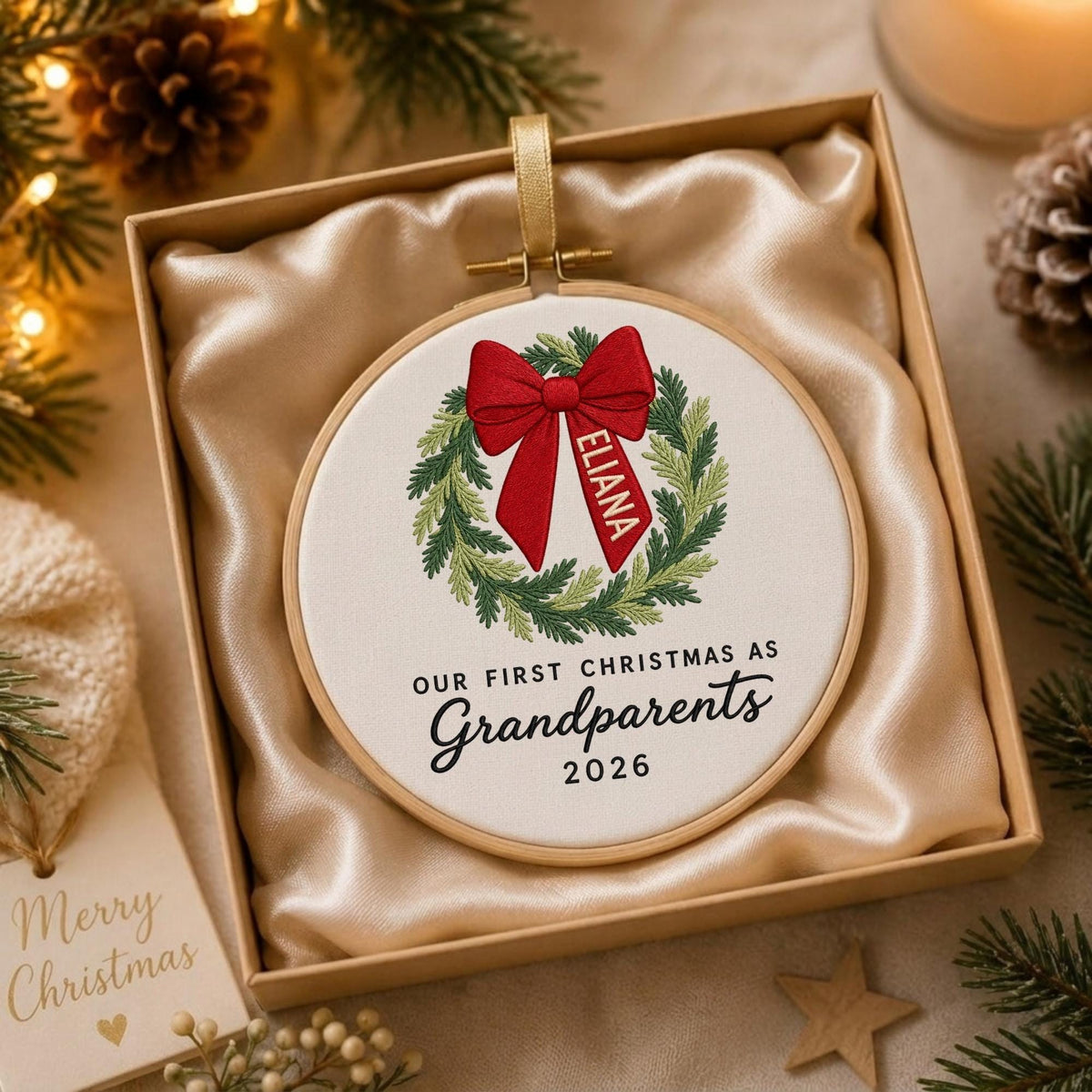 May include: A round wooden embroidery hoop ornament featuring a stitched green wreath with a red bow. The bow has the name ELIANA embroidered on it. Below the wreath, the text reads OUR FIRST CHRISTMAS AS Grandparents 2026 in black.