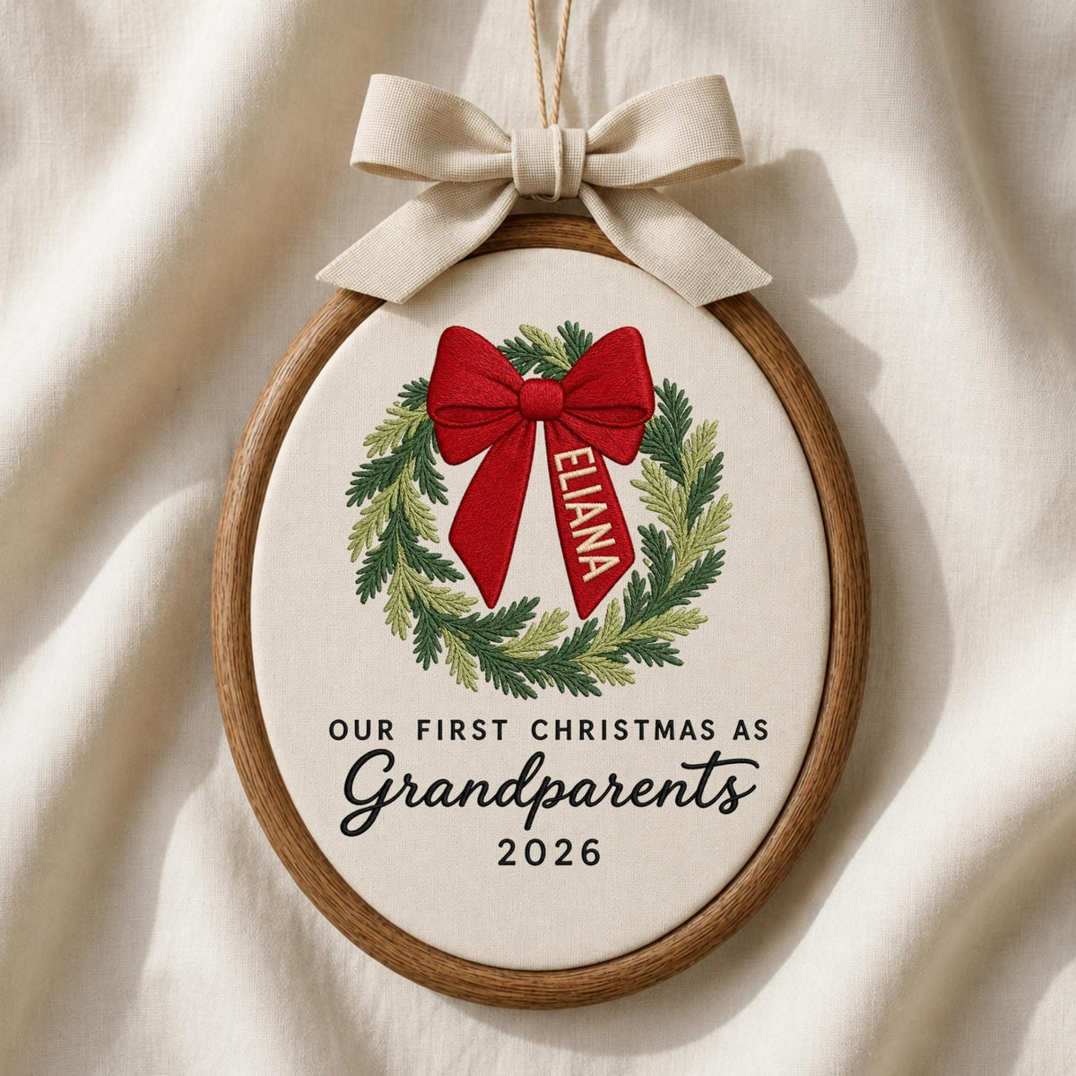 May include: An oval embroidery hoop ornament featuring a green pine wreath with a large red bow. The name ELIANA is embroidered on the bow. Below the wreath, the text reads OUR FIRST CHRISTMAS AS Grandparents 2026. A beige ribbon bow sits on top.