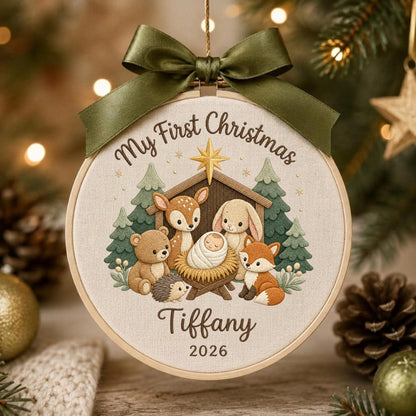 May include: A round embroidery hoop ornament featuring a beige fabric background with a dark green satin bow at the top. The design shows a manger scene with a baby, a deer, a bear, a hedgehog, a rabbit, and a fox. Text reads My First Christmas, Tiffany, and 2026.