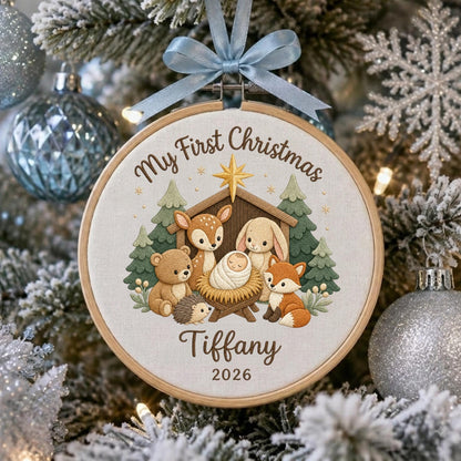 May include: A round wooden embroidery hoop ornament featuring a light fabric background with the text My First Christmas, Tiffany, and 2026. The design shows a manger with a baby, a deer, a bear, a rabbit, a hedgehog, and a fox, topped with a blue ribbon.