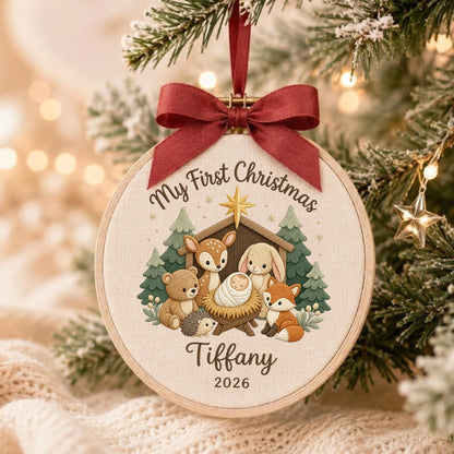 May include: A round wooden embroidery hoop ornament featuring a cream fabric center with a printed illustration of a baby in a manger surrounded by a bear, deer, rabbit, fox, and hedgehog. Text reads My First Christmas, Tiffany, and 2026.