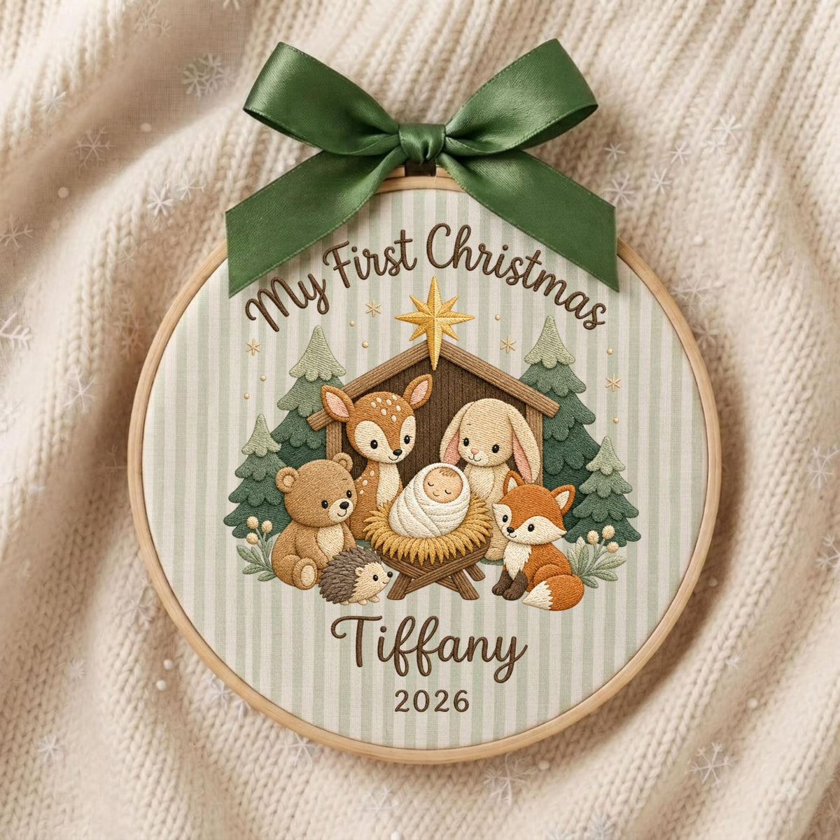 May include: A round wooden embroidery hoop ornament featuring a light green striped fabric background. It displays a printed illustration of a baby in a manger surrounded by a bear, deer, rabbit, hedgehog, and fox. Text reads My First Christmas Tiffany 2026.