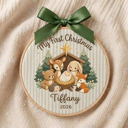 May include: A round wooden embroidery hoop ornament featuring a light green striped fabric background. It displays a printed illustration of a baby in a manger surrounded by a bear, deer, rabbit, hedgehog, and fox. Text reads My First Christmas Tiffany 2026.