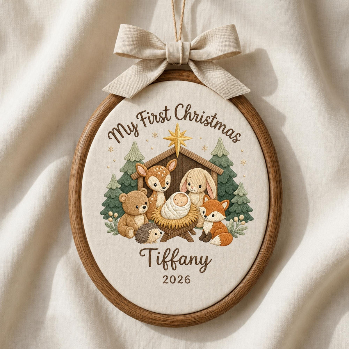 May include: An oval wooden embroidery hoop ornament featuring a cream fabric center with a printed nativity scene of woodland animals, a gold star, and the text My First Christmas, Tiffany, 2026, topped with a beige fabric bow for hanging.