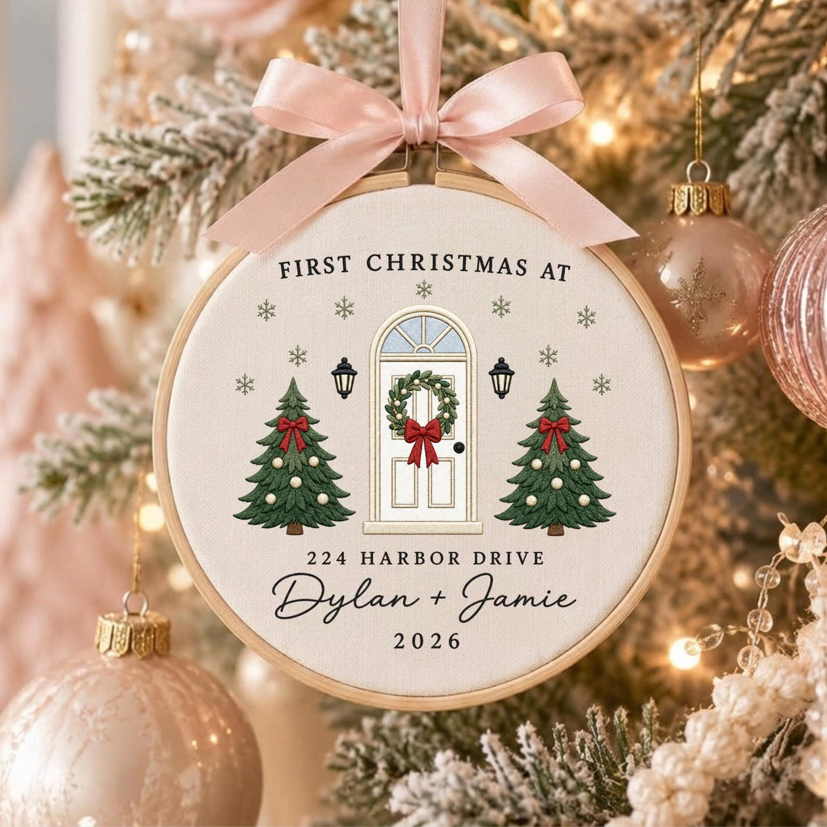 May include: A round embroidery hoop ornament featuring a light beige fabric with a printed design of a white front door, two green Christmas trees, and snowflakes. Text reads: FIRST CHRISTMAS AT 224 HARBOR DRIVE Dylan + Jamie 2026. A pink ribbon bow.