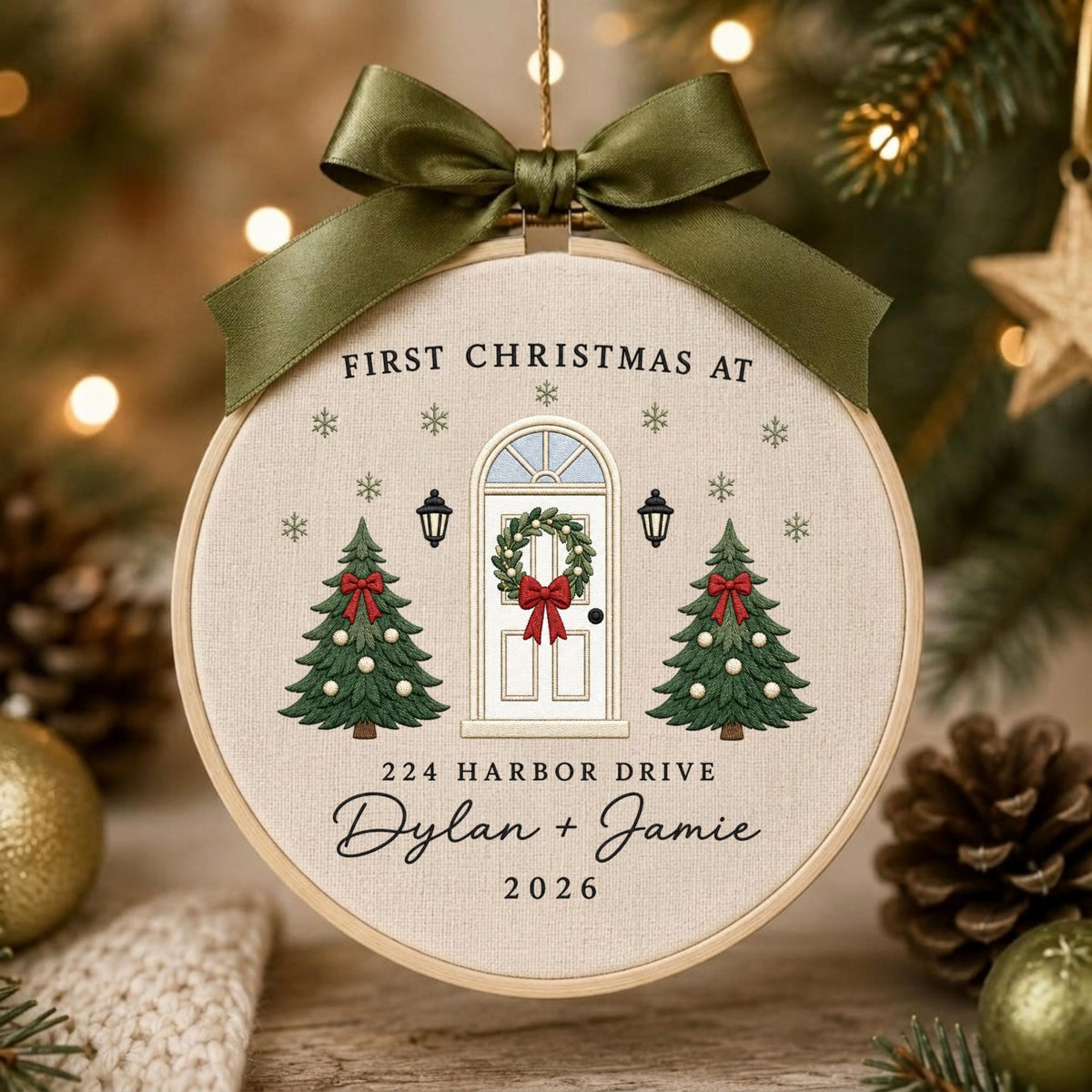 May include: A circular embroidery hoop ornament featuring a cream fabric background with a printed design of a white front door, two green Christmas trees, and snowflakes. Text reads: FIRST CHRISTMAS AT 224 HARBOR DRIVE Dylan + Jamie 2026.