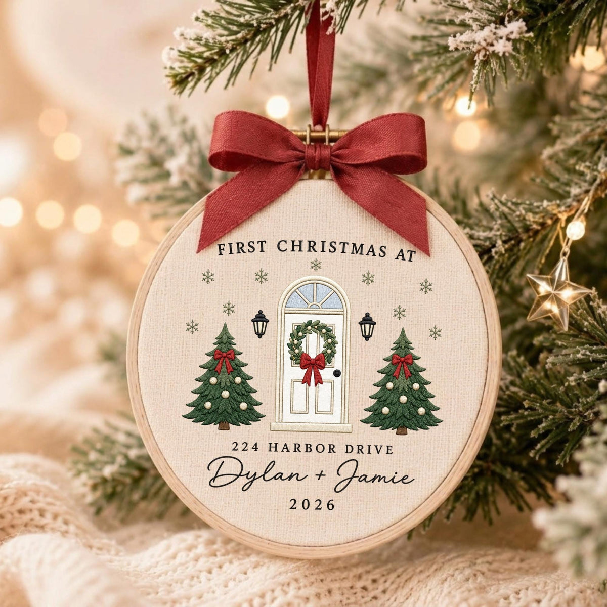 May include: A round embroidery hoop ornament featuring a cream fabric background with a printed design of a front door, two Christmas trees, and snowflakes. Text reads: FIRST CHRISTMAS AT 224 HARBOR DRIVE Dylan + Jamie 2026. A red ribbon bow is attached.