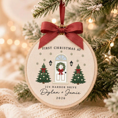 May include: A round embroidery hoop ornament featuring a cream fabric background with a printed design of a front door, two Christmas trees, and snowflakes. Text reads: FIRST CHRISTMAS AT 224 HARBOR DRIVE Dylan + Jamie 2026. A red ribbon bow is attached.