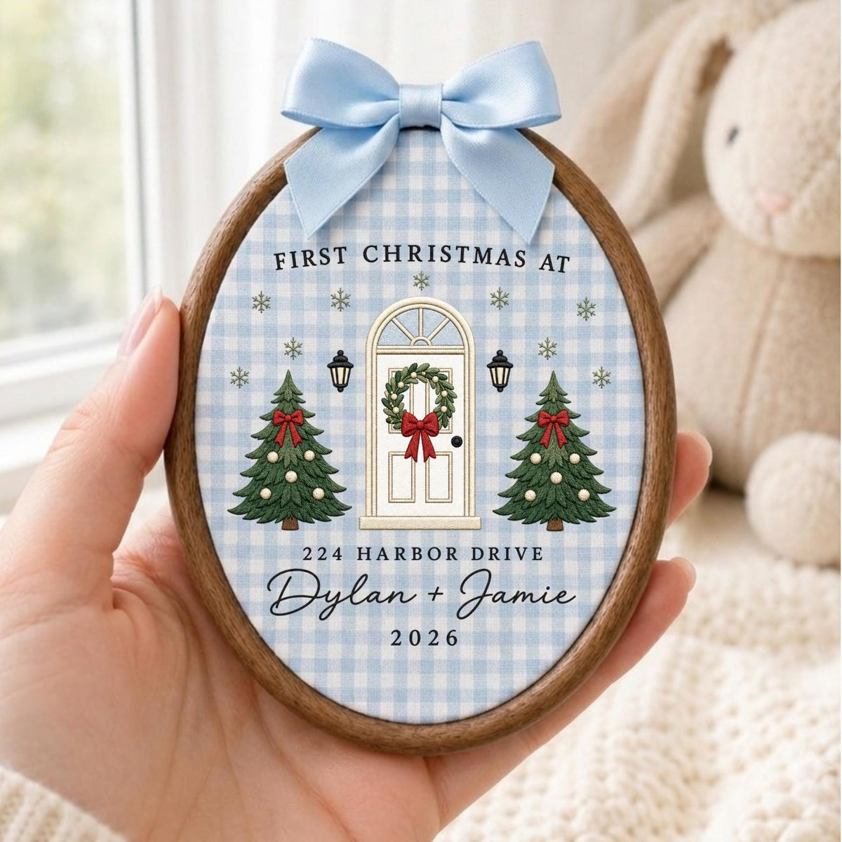May include: An oval wooden embroidery hoop ornament featuring a light blue gingham fabric background. The design shows a white front door with a wreath, two green Christmas trees, and the text: FIRST CHRISTMAS AT 224 HARBOR DRIVE Dylan + Jamie 2026. A light blue satin bow is attached to the top.
