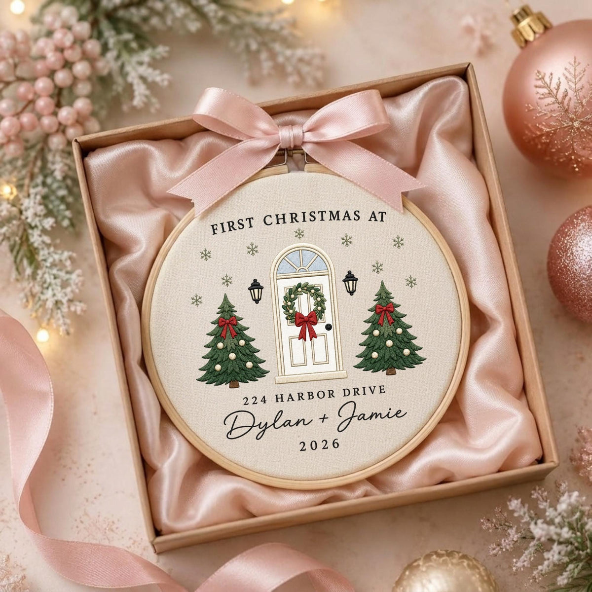 May include: A round wooden embroidery hoop featuring a cream fabric background with a stitched design of a front door, two Christmas trees, and snowflakes. Text reads: FIRST CHRISTMAS AT 224 HARBOR DRIVE Dylan + Jamie 2026. A pink satin bow is attached.
