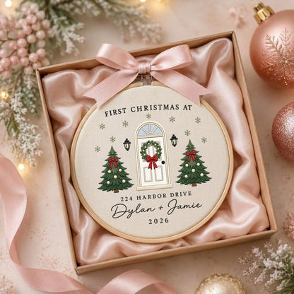 May include: A round wooden embroidery hoop featuring a cream fabric background with a stitched design of a front door, two Christmas trees, and snowflakes. Text reads: FIRST CHRISTMAS AT 224 HARBOR DRIVE Dylan + Jamie 2026. A pink satin bow is attached.