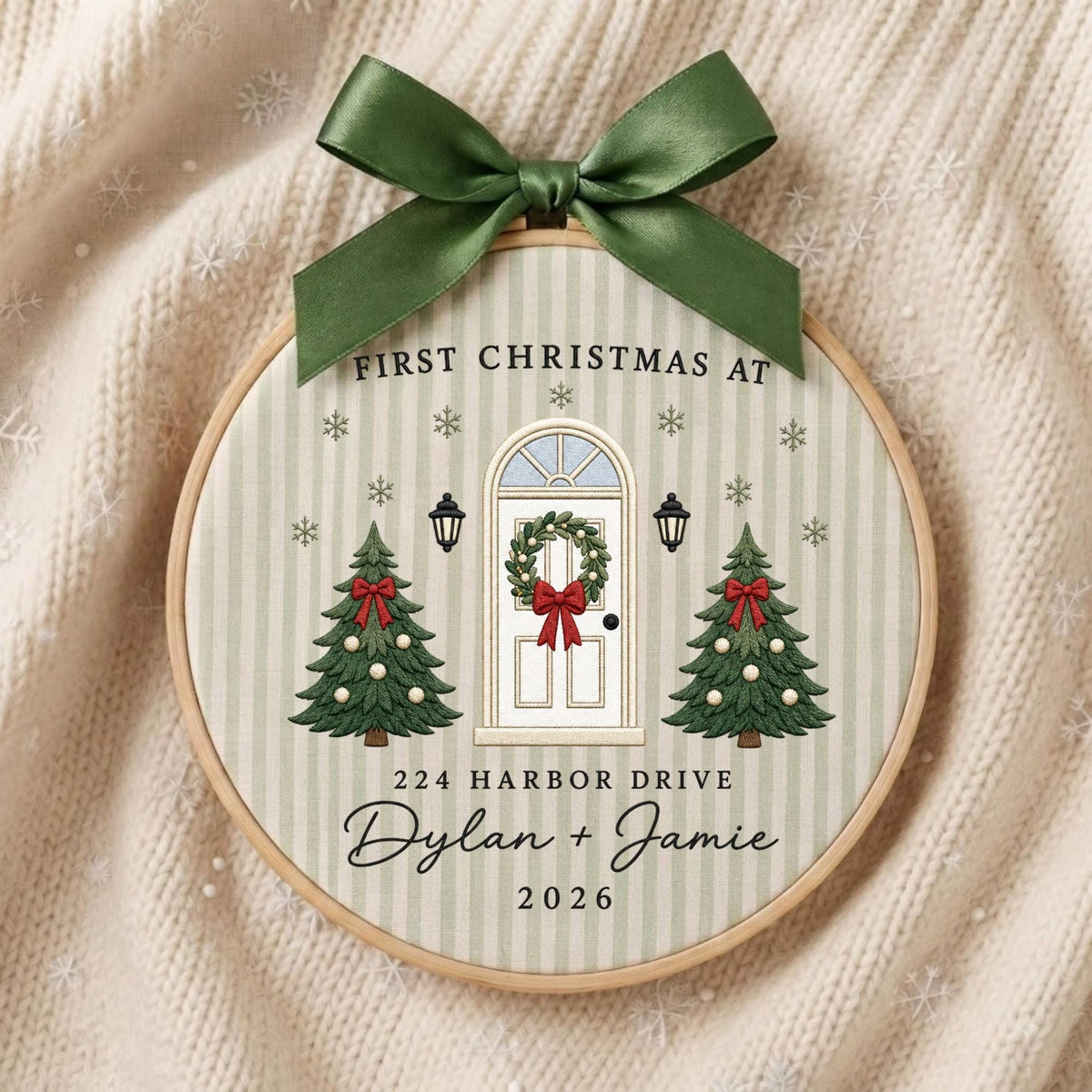 May include: A round embroidery hoop ornament featuring a light green striped fabric with a printed design of a white front door, two Christmas trees, and snowflakes. It includes the text FIRST CHRISTMAS AT 224 HARBOR DRIVE Dylan + Jamie 2026.