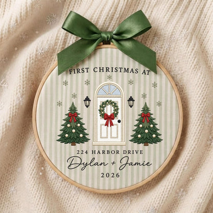 May include: A round embroidery hoop ornament featuring a light green striped fabric with a printed design of a white front door, two Christmas trees, and snowflakes. It includes the text FIRST CHRISTMAS AT 224 HARBOR DRIVE Dylan + Jamie 2026.