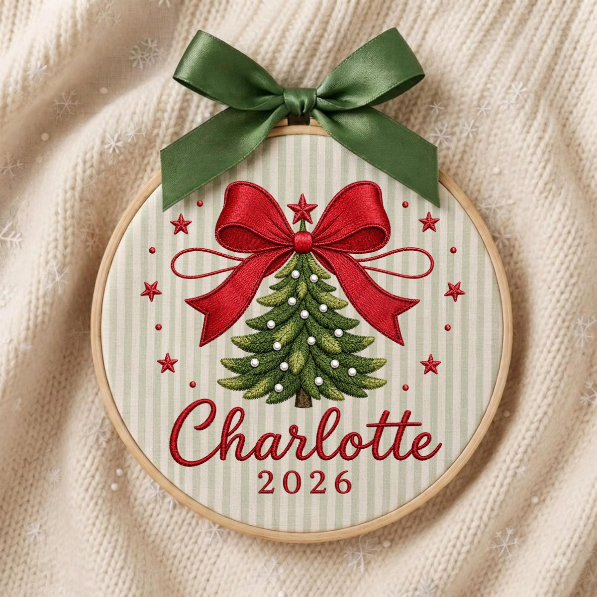 May include: A circular wooden embroidery hoop featuring a light green and white striped fabric background. The design includes an embroidered green Christmas tree with white accents, a large red bow, red stars, and the text Charlotte 2026.
