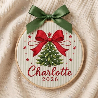 May include: A circular wooden embroidery hoop featuring a light green and white striped fabric background. The design includes an embroidered green Christmas tree with white accents, a large red bow, red stars, and the text Charlotte 2026.