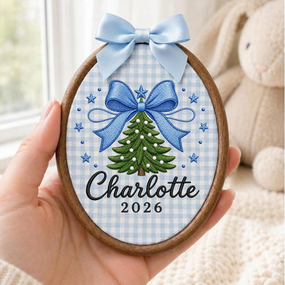 May include: An oval wooden embroidery hoop ornament featuring a light blue and white gingham fabric background. The design includes an embroidered green Christmas tree with white accents, a large blue bow, stars, and the text Charlotte 2026.