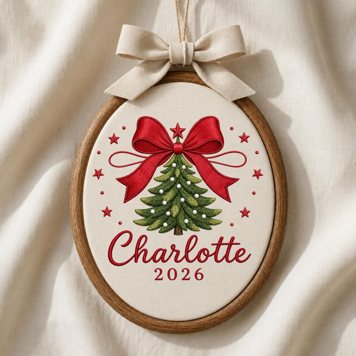 May include: An oval wooden embroidery hoop containing a cream-colored fabric with a stitched green Christmas tree, a large red bow, and red stars. The text Charlotte and 2026 are embroidered in red. A beige fabric bow is attached to the top.