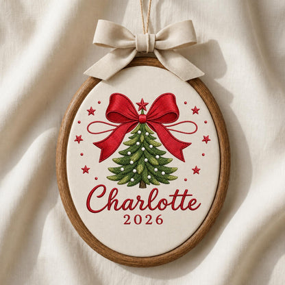 May include: An oval wooden embroidery hoop containing a cream-colored fabric with a stitched green Christmas tree, a large red bow, and red stars. The text Charlotte and 2026 are embroidered in red. A beige fabric bow is attached to the top.