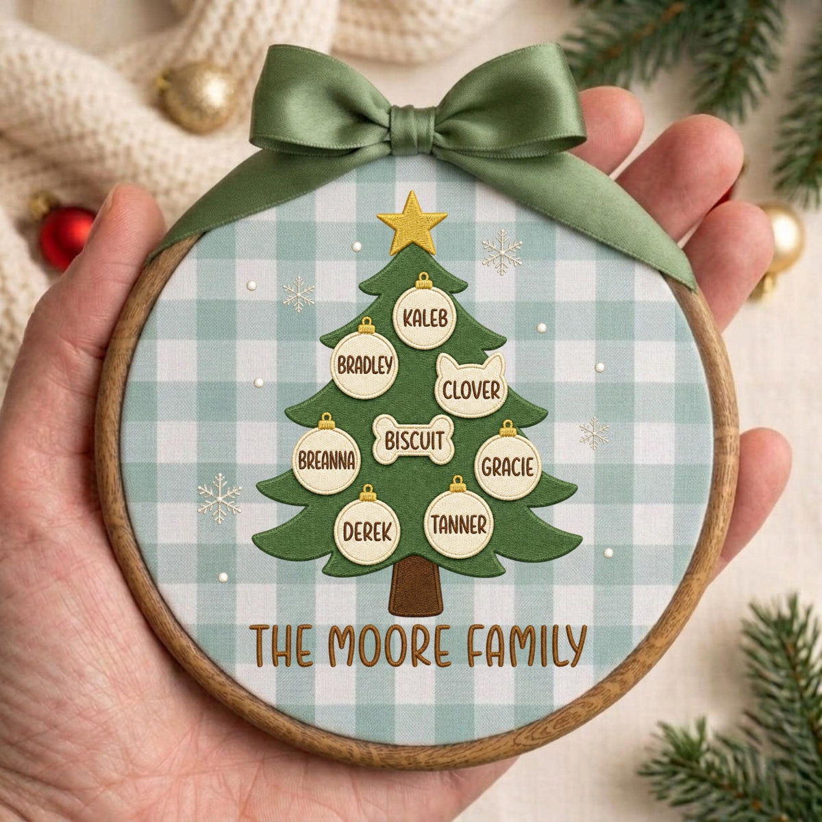 May include: A round wooden embroidery hoop ornament featuring a light blue and white gingham fabric background. A green Christmas tree illustration is centered with a gold star, white ornaments, and the text THE MOORE FAMILY in brown.