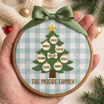 May include: A round wooden embroidery hoop ornament featuring a light blue and white gingham fabric background. A green Christmas tree illustration is centered with a gold star, white ornaments, and the text THE MOORE FAMILY in brown.