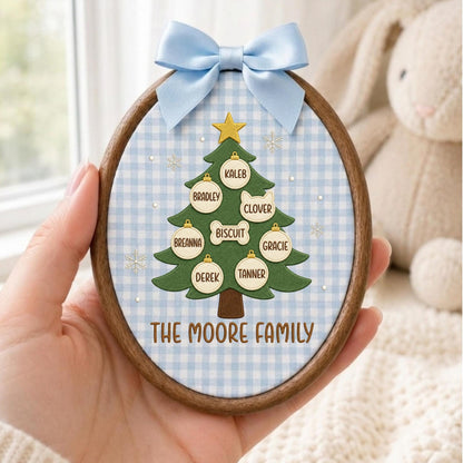 May include: An oval wooden embroidery hoop frame containing a light blue and white gingham fabric background. A green Christmas tree illustration features a gold star, white snowflake accents, and ornaments labeled with names and pet names.