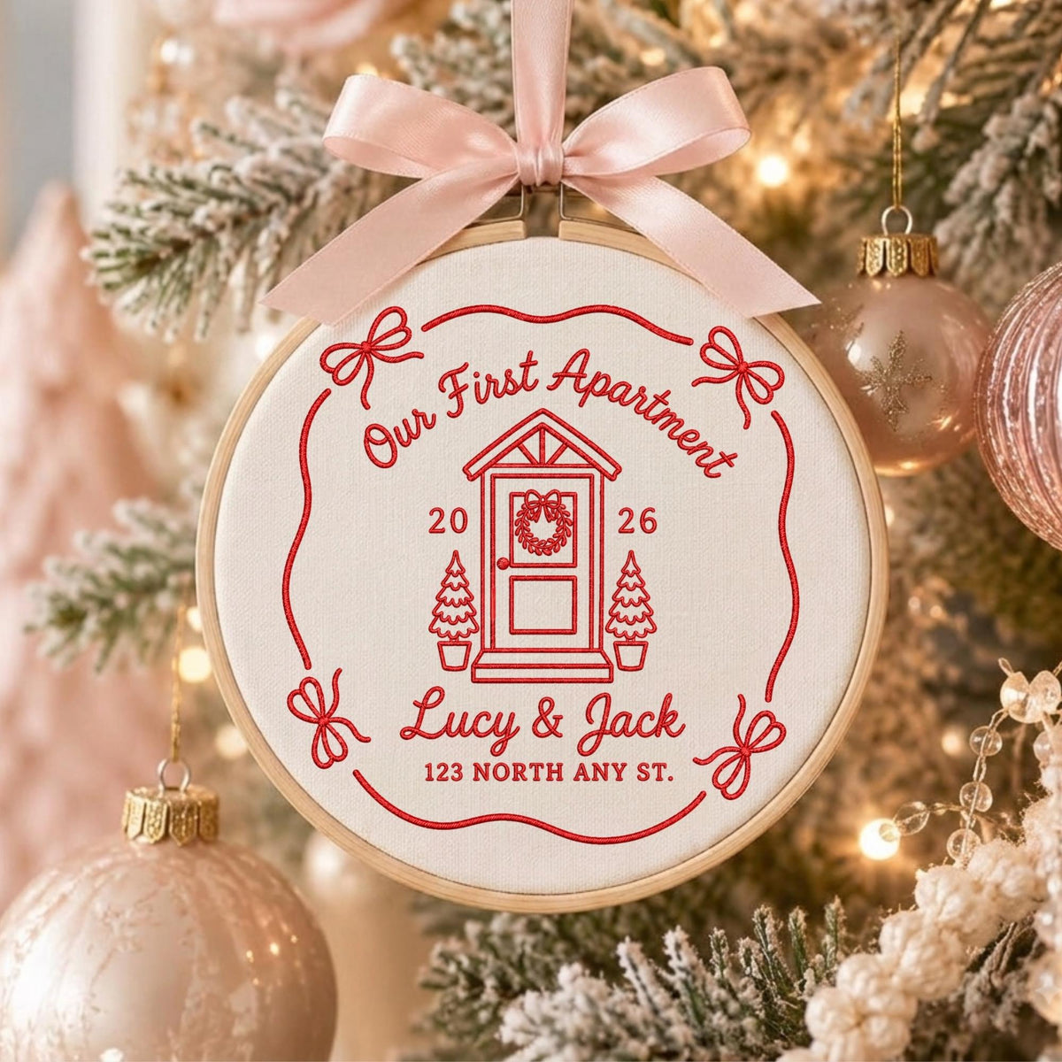 May include: A round embroidery hoop ornament featuring cream fabric with red stitched text and graphics. The design includes a house door, two small trees, and the text Our First Apartment, 2026, Lucy &amp; Jack, and 123 NORTH ANY ST. A light pink ribbon bow is tied at the top.