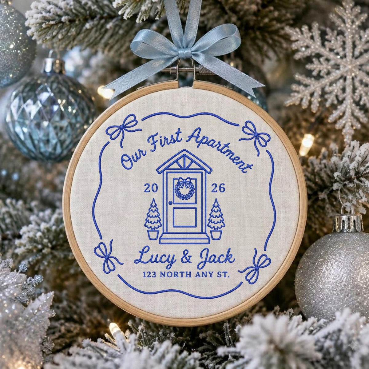 May include: A circular wooden embroidery hoop containing off-white fabric with blue stitched text and graphics. The design features a house door, two small trees, bows, and the text Our First Apartment, 2026, Lucy &amp; Jack, 123 NORTH ANY ST.