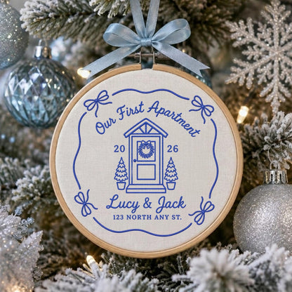 May include: A circular wooden embroidery hoop containing off-white fabric with blue stitched text and graphics. The design features a house door, two small trees, bows, and the text Our First Apartment, 2026, Lucy &amp; Jack, 123 NORTH ANY ST.
