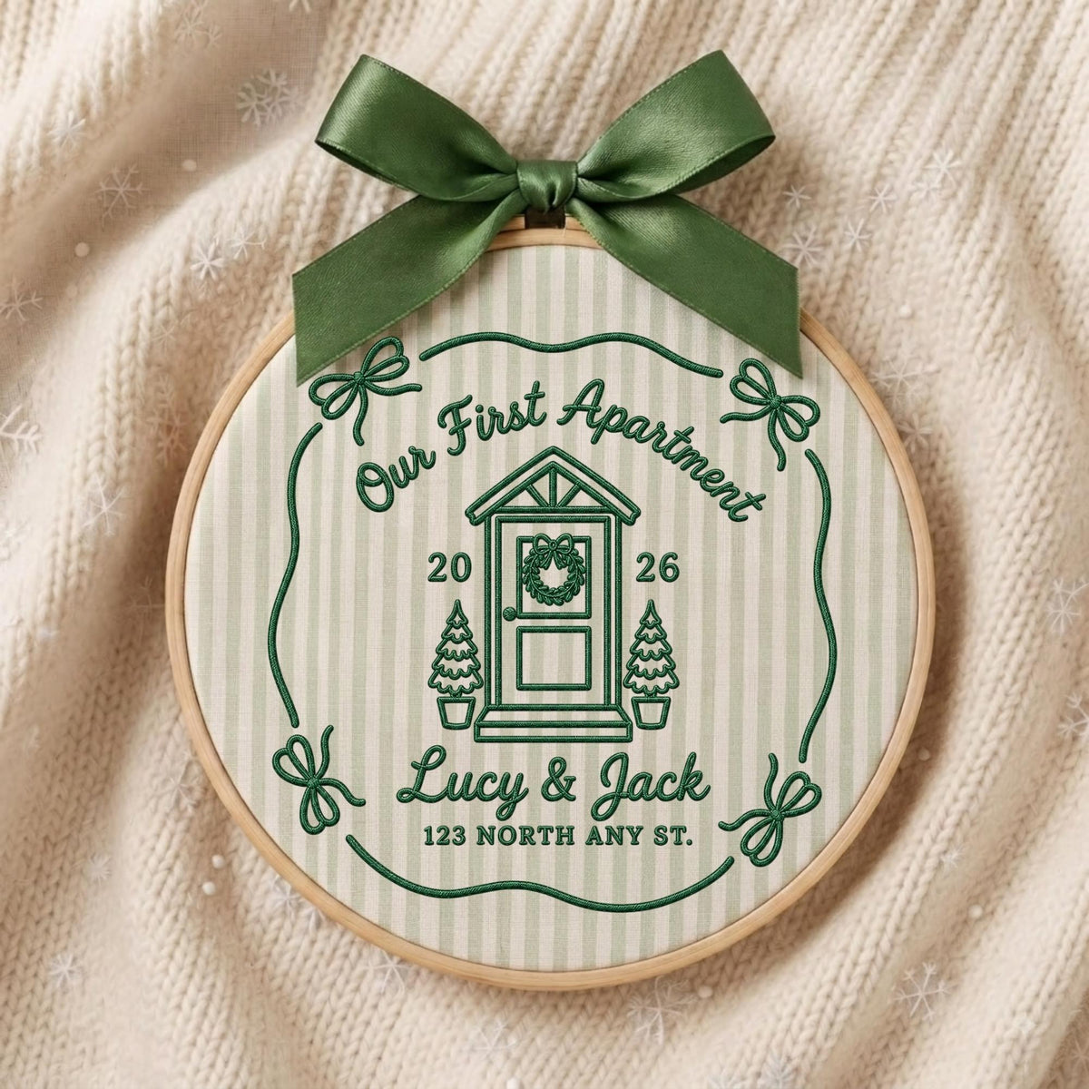 May include: A circular embroidery hoop featuring a light green and white striped fabric with dark green embroidery. The design includes a house illustration, the text "Our First Apartment", "2026", "Lucy &amp; Jack", and "123 NORTH ANY ST.", topped with a dark green satin bow.